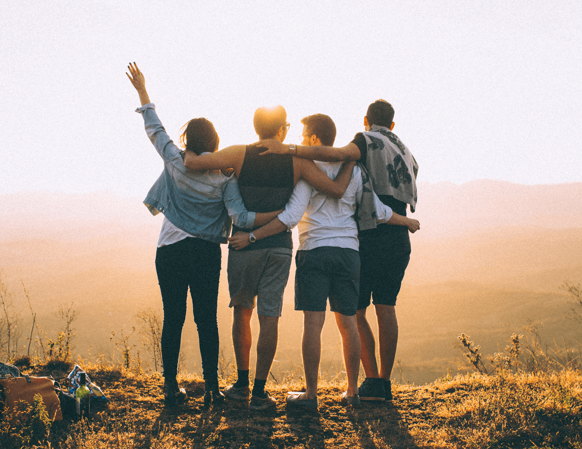 The back of four friends standing side-by-side, embracing one another, arms outstretched, enjoying the view of the horizon.
