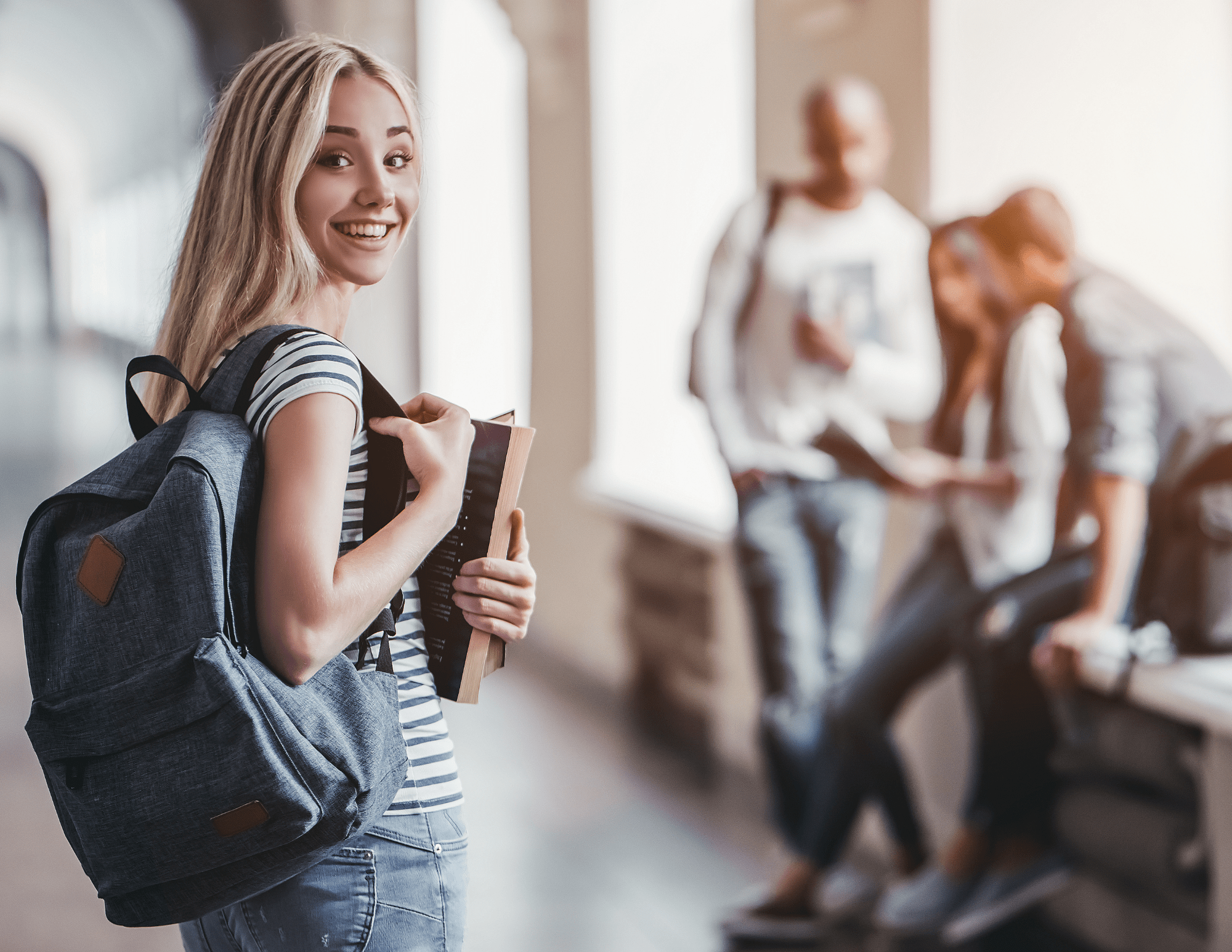 A smiling young blond woman with a backpack and textbook, standing in a school hallway. Three out-of-focus students sit in the background.