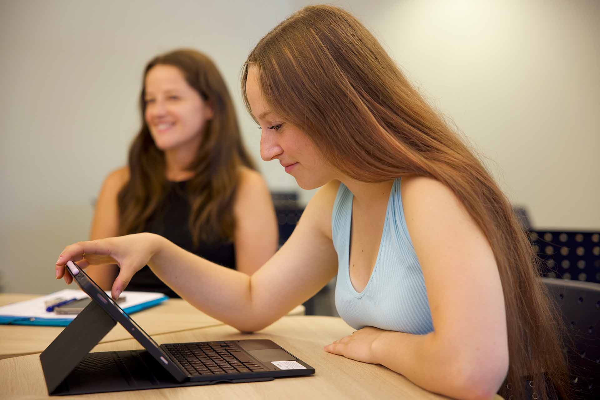2 – ExploreProgramPage_Image10 – FINAL_ A teenager wearing headphones writes in a notebook while looking at an open laptop in a study room or office setting.