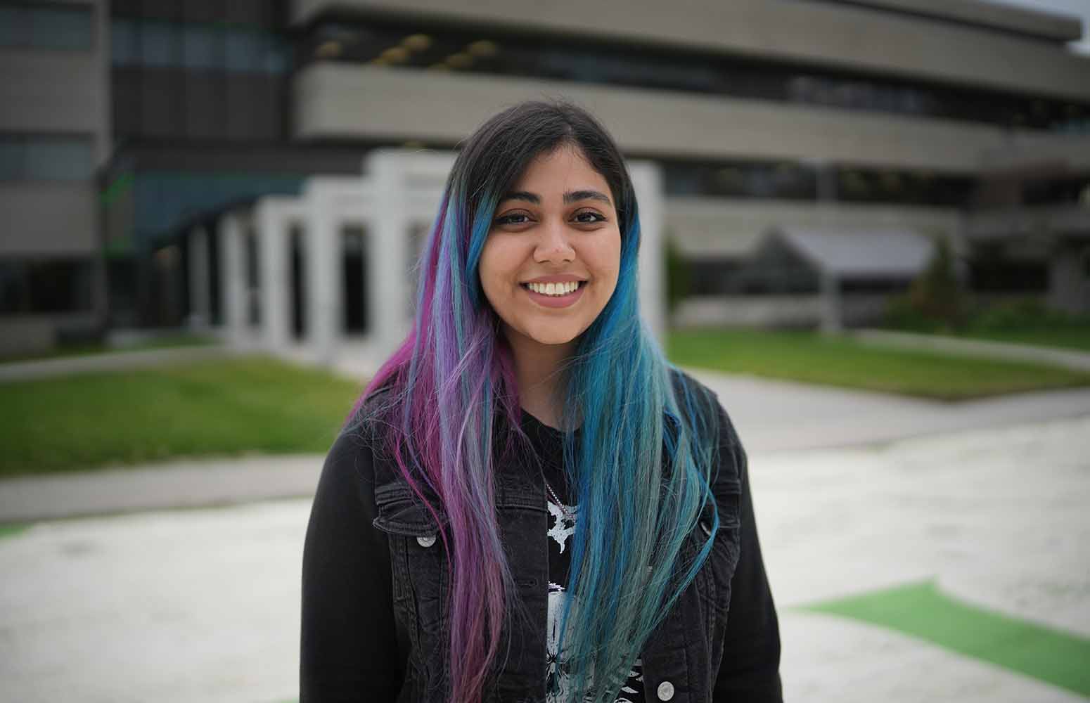 A smiling young woman with colorful hair standing outdoors in front of a building.