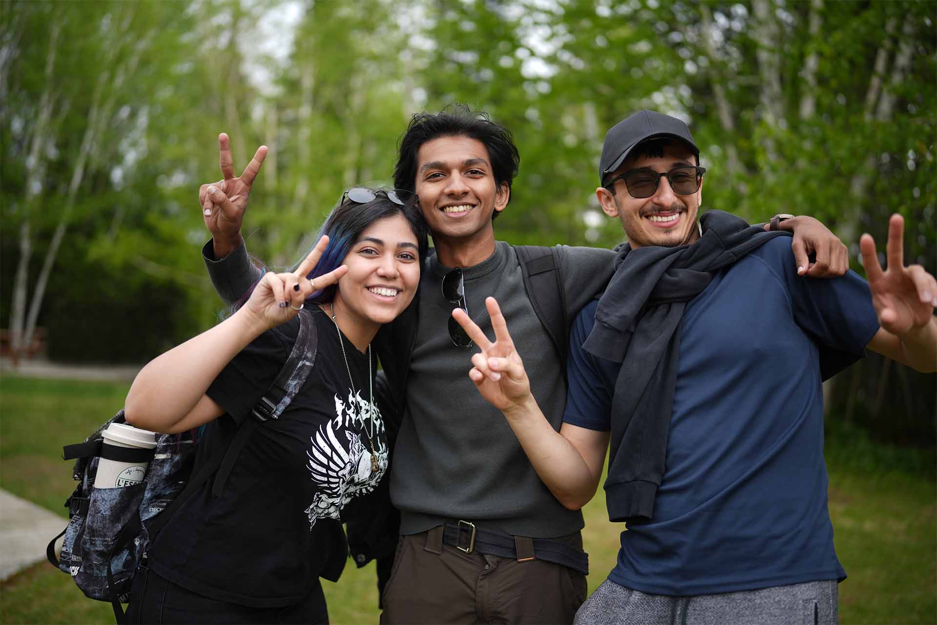 2 – ExploreProgramPage_Image9 – FINAL (EDITED) Three smiling young adults wearing backpacks pose together outdoors on a forest trail, making peace signs with their hands while enjoying a nature outing.