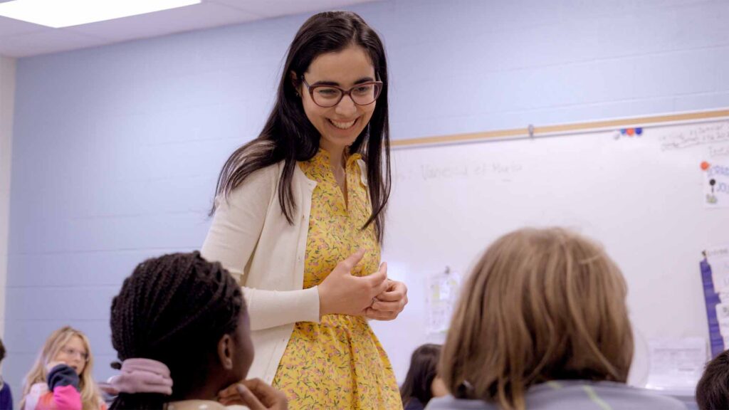 Students sitting at a classroom desk while their teacher leans over, smiling and pointing at something on a tablet, during a group activity.
