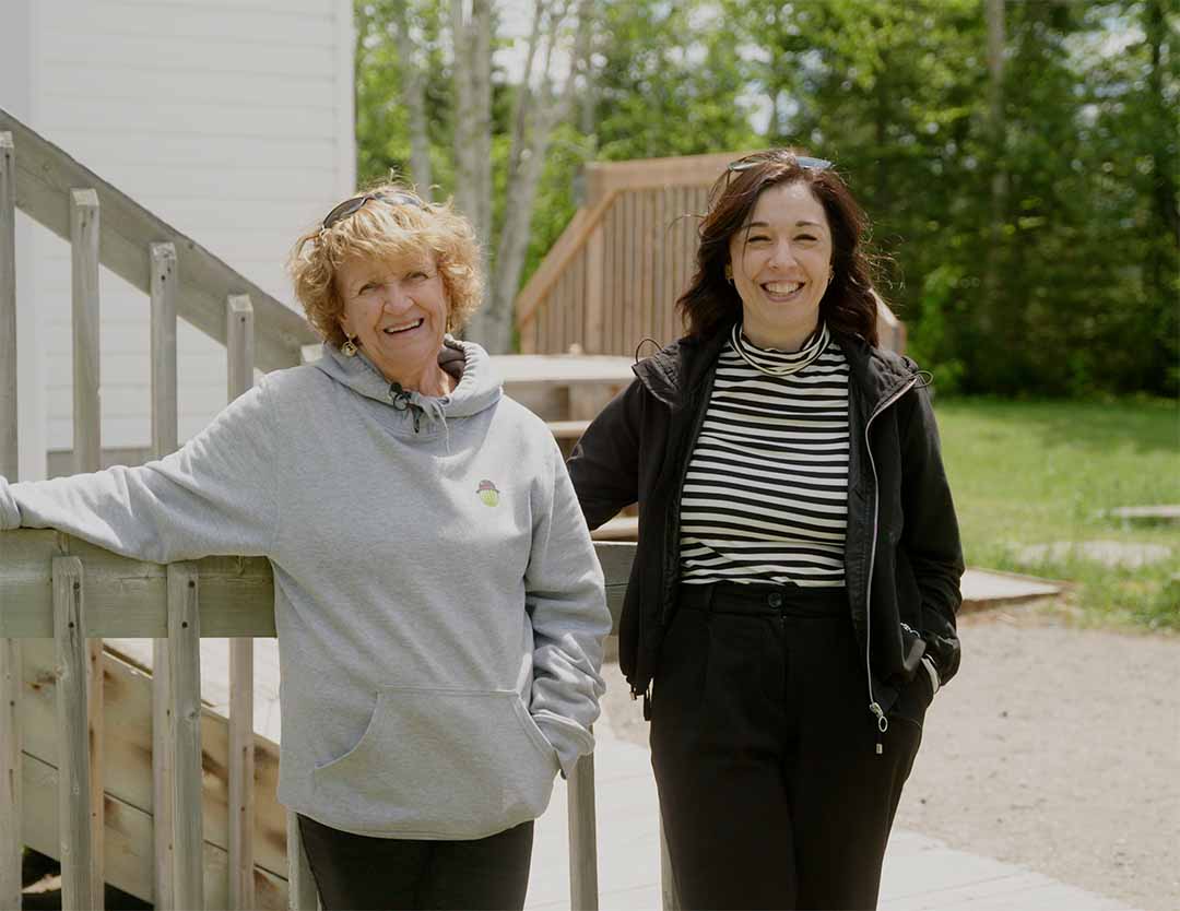 Two women stand outdoors beside an accessible wooden ramp leading to a white building. The women are smiling at the camera with trees and a grassy area in the background.