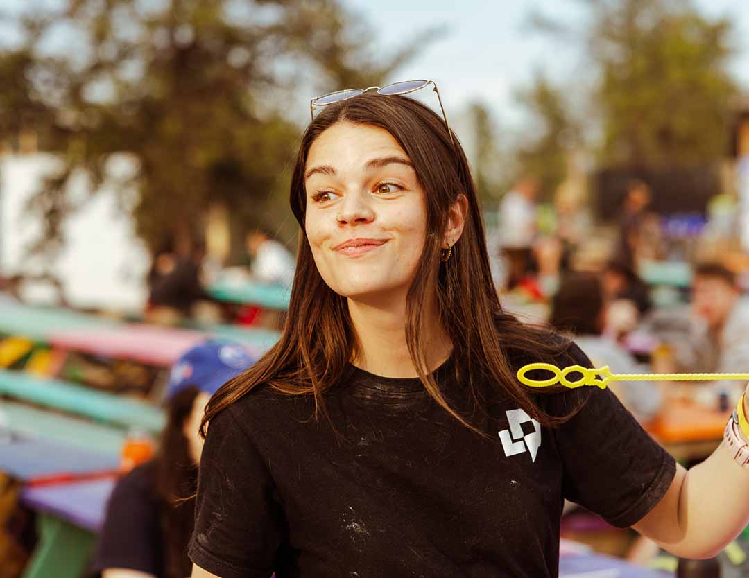 A young woman holds a bubble wand at an outdoor gathering, with colourful picnic tables and a blurred crowd in the background.
