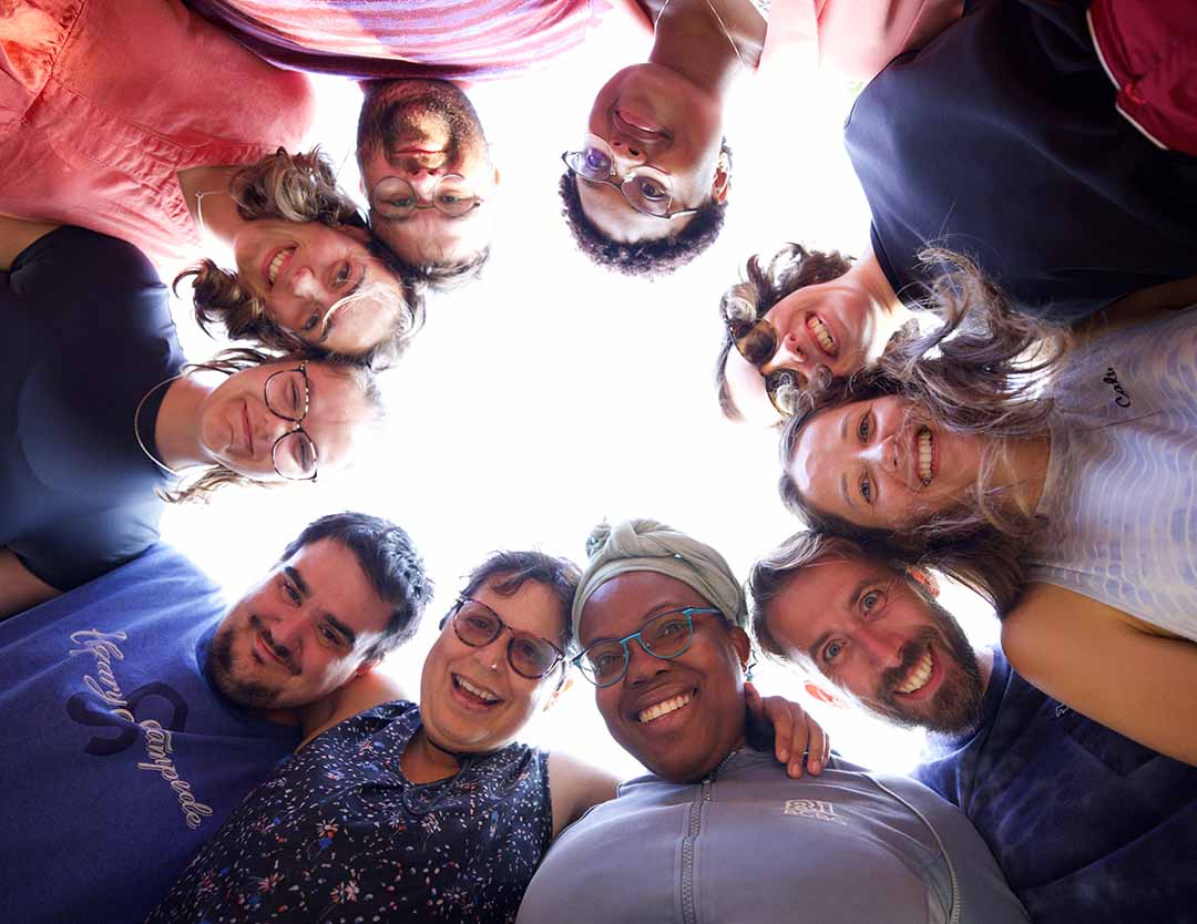A group of ten people stand in a circle outdoors, looking down toward the camera and smiling, with the bright sky visible in the centre of the group.