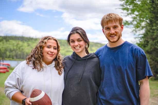 Three youth smiling and standing close together outdoors, with trees, hills, and canoes by a lake in the background; the person on the left holds an American football.