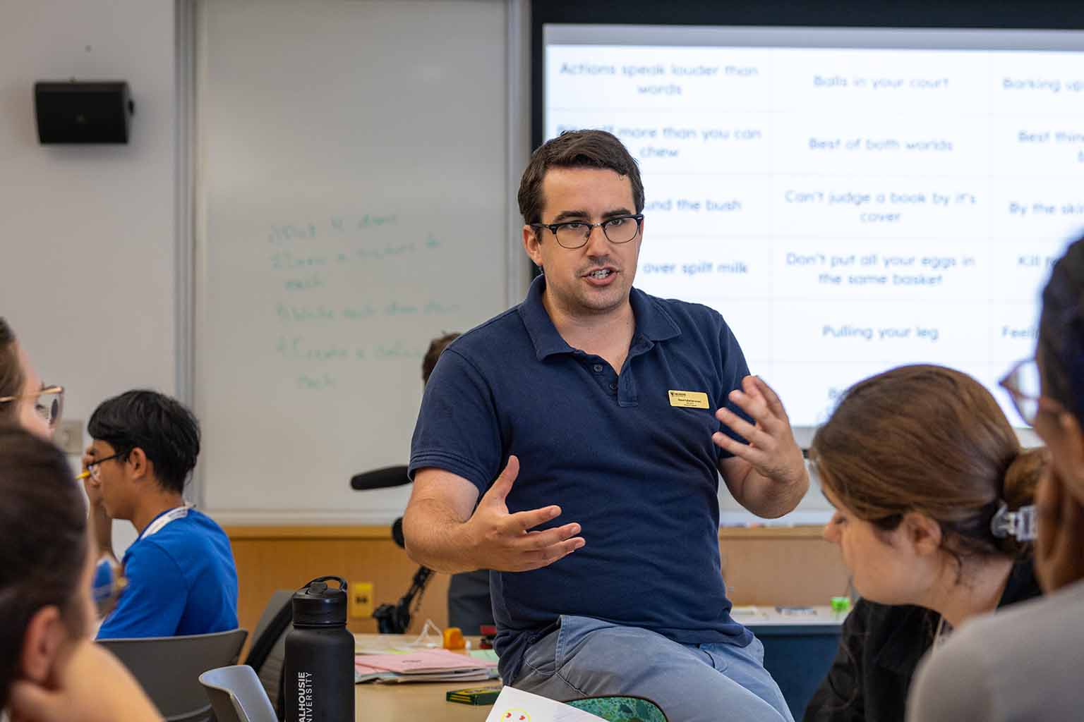 An Odyssey Language Assistant sits on a desk at the front of a classroom, gesturing as he speaks to a small group of students gathered around a table, with a whiteboard and projected slide behind him.