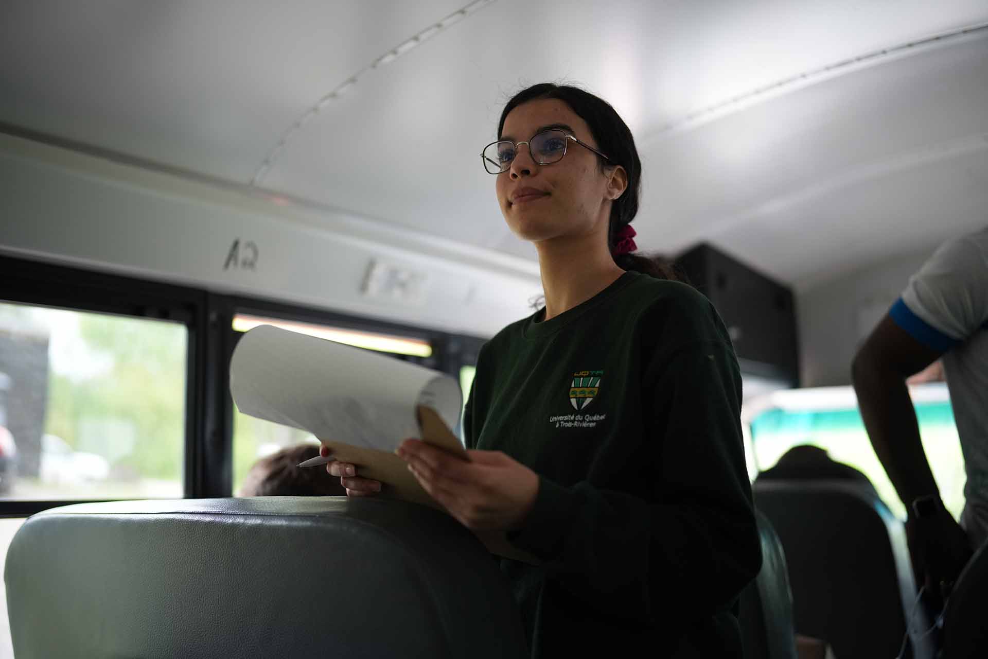 6 – GetInvolvedPage_Image22 – FINAL A young woman standing inside a school bus holding papers while speaking to other passengers during an educational outing.