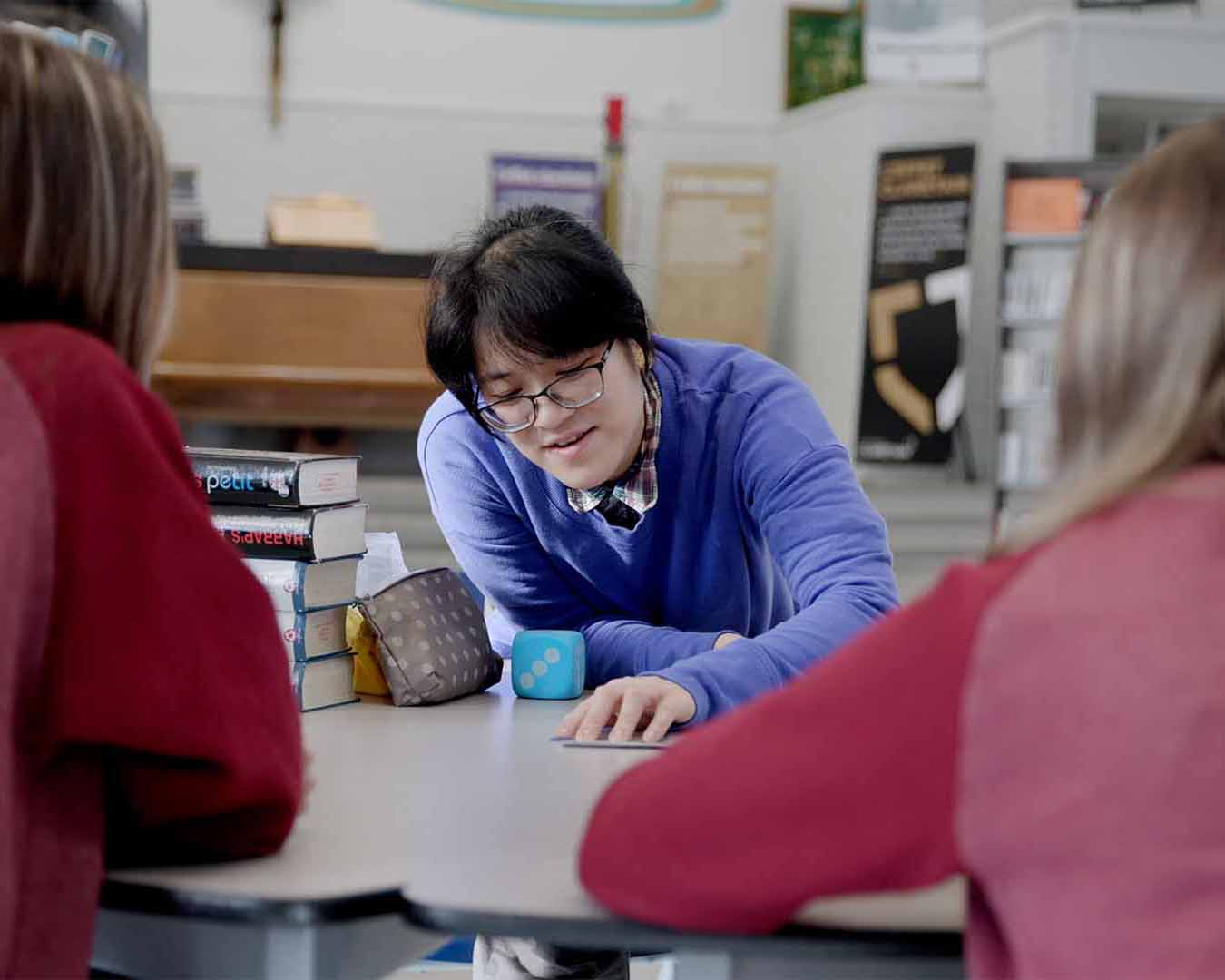 A language assistant leans over a table to demonstrate an activity for students in a bright classroom, with books and learning materials visible on the table. The students are facing the assistant and have their backs to the camera.