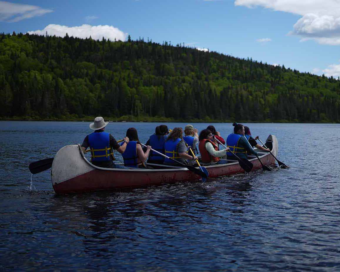 Nine people wearing life jackets are paddling a canoe across a calm lake and facing away from the camera. The group is surrounded by forested hills under a bright blue sky with scattered clouds.