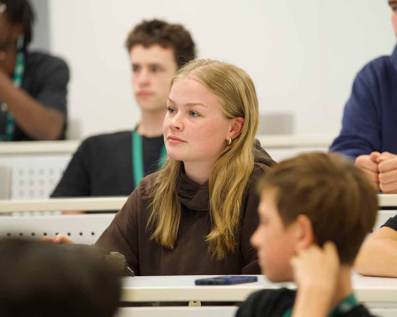 A student listens attentively during a classroom session while other participants are seated in the background, engaged in a group learning environment.